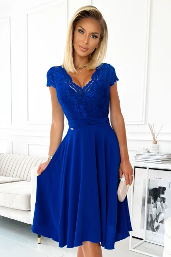 Kobieta na zdjęciu prezentuje elegancką sukienkę wieczorową w głębokim odcieniu niebieskiego, która doskonale podkreśla jej sylwetkę. Sukienka wieczorowa z koronkowym wykończeniem i delikatnym dekoltem dodaje całości wyjątkowego szyku i klasy.