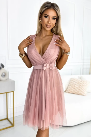 Modelka prezentuje elegancką sukienkę tulle w odcieniu pudrowego różu, która subtelnie podkreśla kobiecą sylwetkę. Sukienka tulle wyróżnia się delikatnym materiałem i uroczym wiązaniem w talii, dodającym całości romantycznego charakteru.