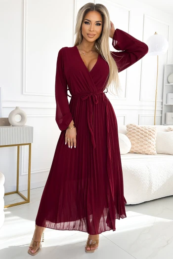 Modelka prezentuje elegancką sukienkę maxi w głębokim odcieniu bordo, która idealnie podkreśla sylwetkę dzięki wiązaniu w talii. Sukienka maxi z długimi rękawami i delikatnym plisowaniem dodaje klasy i szyku każdej stylizacji.