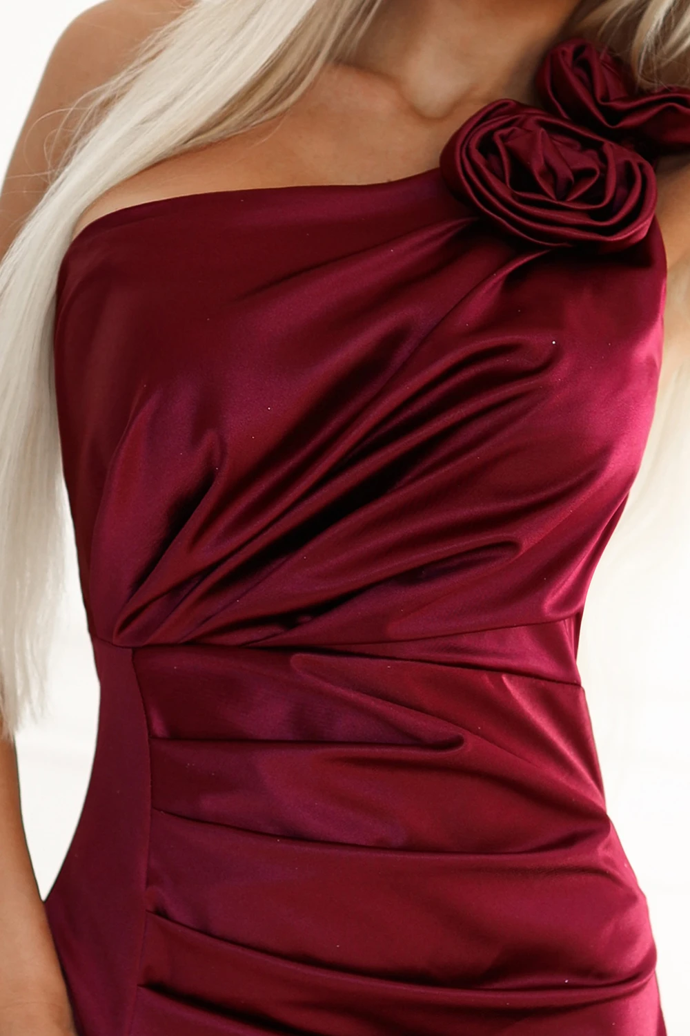 Kobieta na zdjęciu prezentuje elegancką sukienkę wieczorową w głębokim odcieniu burgundu, która przyciąga uwagę dzięki efektownemu marszczeniu i ozdobnym kwiatom na ramieniu. Sukienka wieczorowa idealnie podkreśla sylwetkę, dodając jej szyku i klasy.