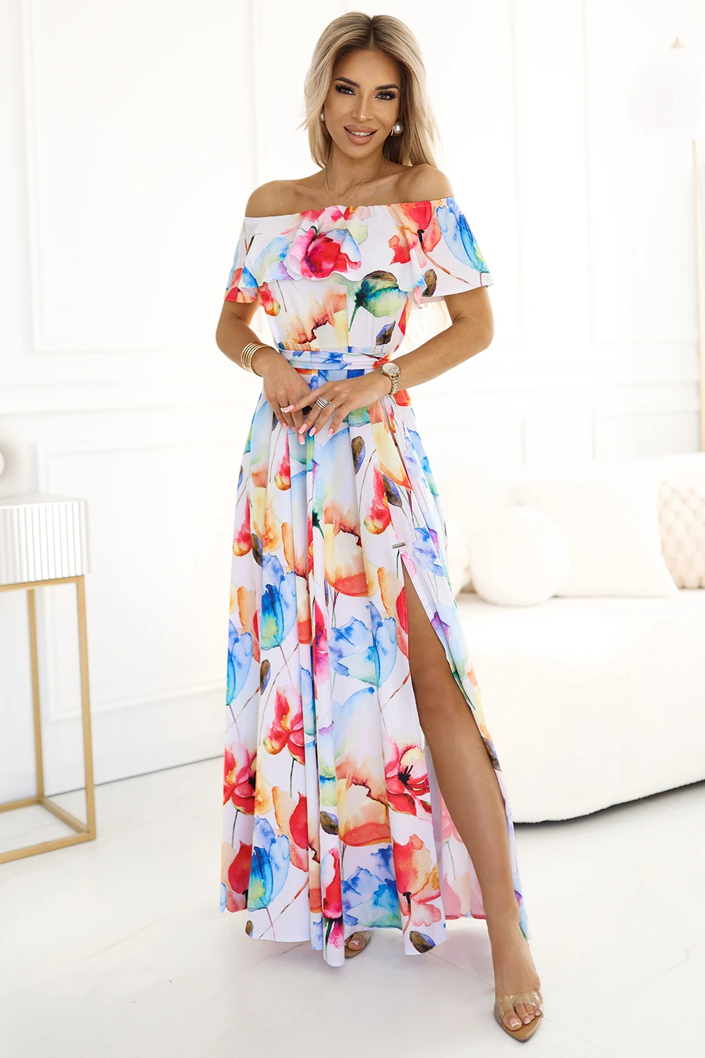 Modelka prezentuje elegancką sukienkę maxi w kwiaty, która zachwyca swoją kolorystyką i zwiewnym fasonem. Sukienka maxi w kwiaty idealnie podkreśla kobiecą sylwetkę, a odkryte ramiona dodają jej subtelnego uroku.