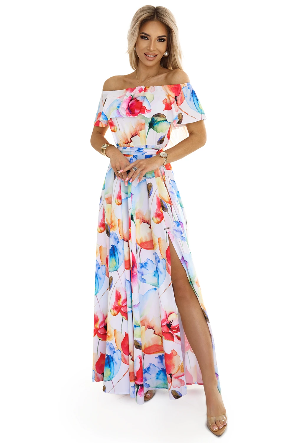 Kobieta na zdjęciu prezentuje elegancką sukienkę maxi w kwiaty, która przyciąga wzrok swoim kolorowym wzorem. Sukienka maxi w kwiaty z odkrytymi ramionami i subtelnym rozcięciem dodaje stylizacji lekkości i kobiecego uroku.
