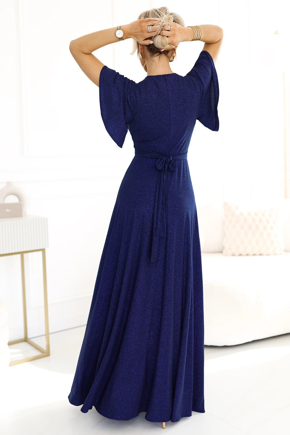 Modelka prezentuje elegancką, granatową sukienkę maxi z delikatnym połyskiem, która idealnie podkreśla sylwetkę. Sukienka maxi z rozkloszowanym dołem i wiązaniem w talii dodaje szyku i klasy każdej stylizacji.
