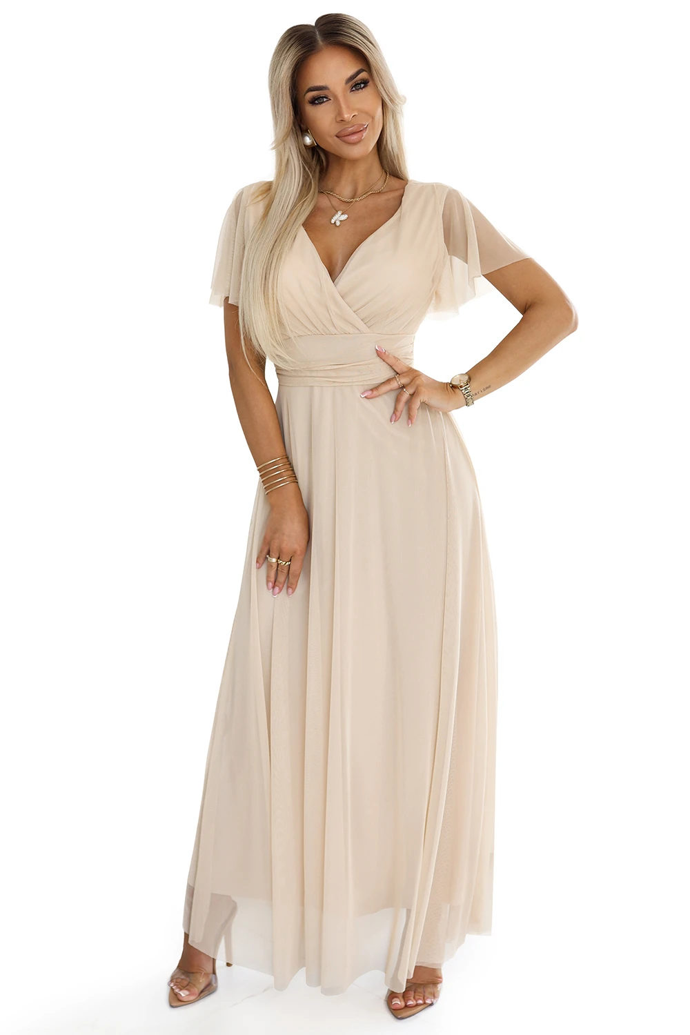 Modelka prezentuje elegancką sukienkę maxi, która zachwyca delikatnym beżowym odcieniem i zwiewnym materiałem. Sukienka maxi podkreśla kobiecą sylwetkę dzięki subtelnemu dekoltowi i dopasowanej talii, idealna na wyjątkowe okazje.