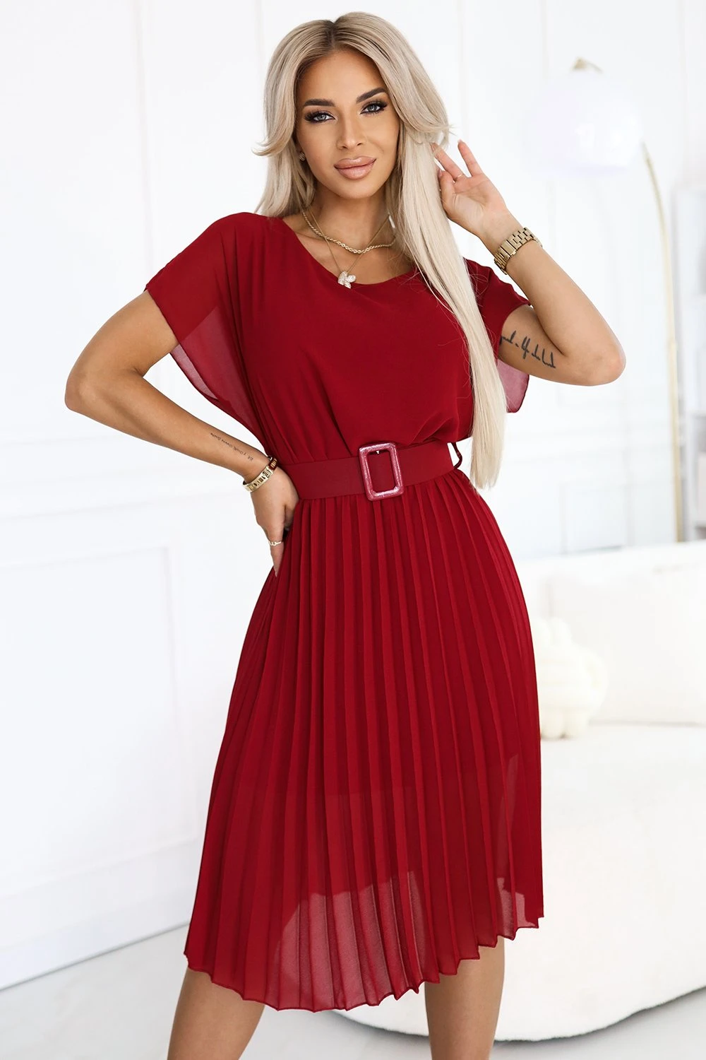 Modelka prezentuje elegancką czerwoną sukienkę z plisowanym dołem, która idealnie podkreśla kobiecą sylwetkę. Sukienka z paskiem w talii dodaje stylizacji wyrafinowanego charakteru, czyniąc ją doskonałym wyborem na wyjątkowe okazje.