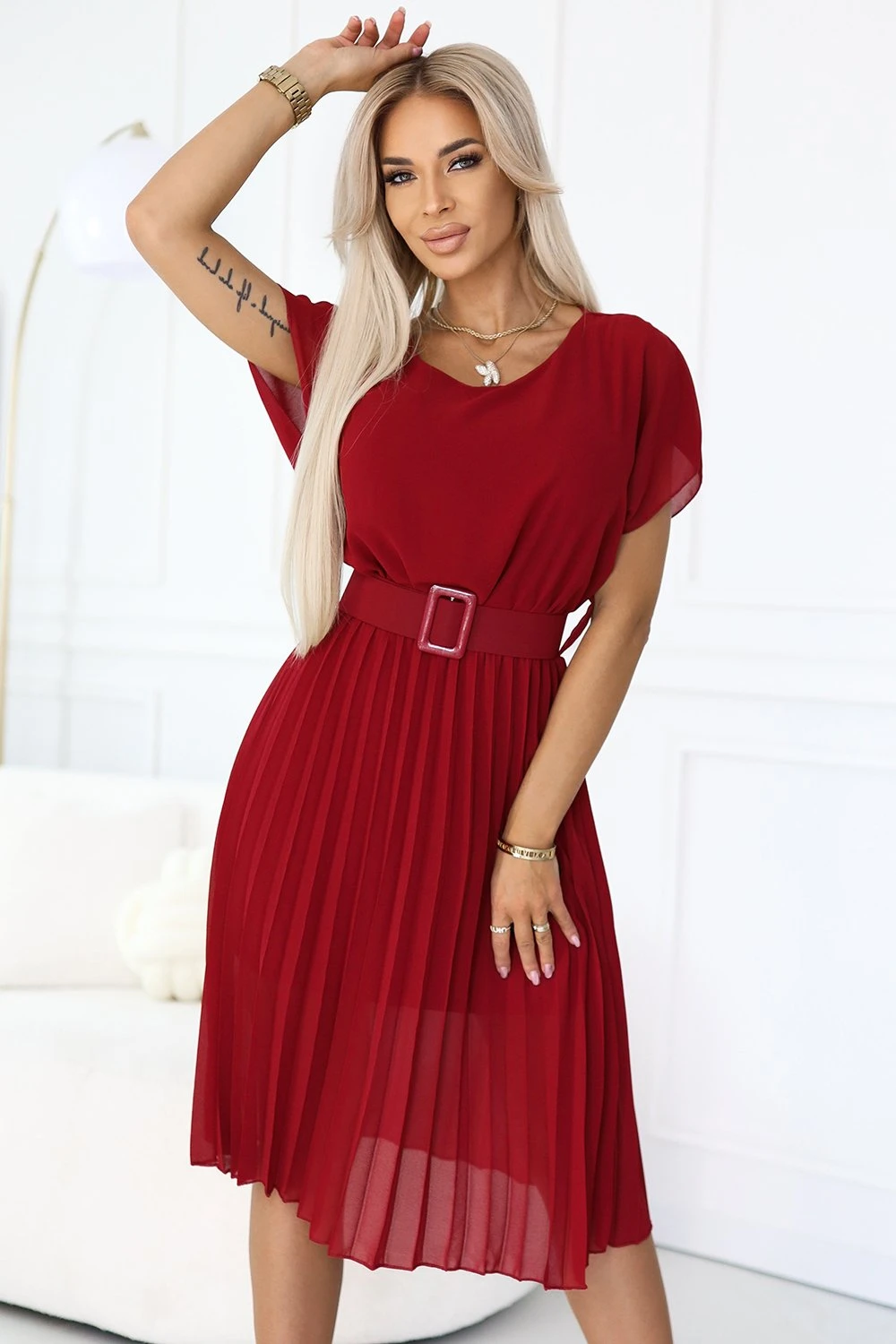 Kobieta na zdjęciu prezentuje elegancką sukienkę z paskiem, która podkreśla jej sylwetkę i dodaje szyku. Sukienka z paskiem wyróżnia się głębokim czerwonym kolorem i plisowanym dołem, idealnym na różne okazje.