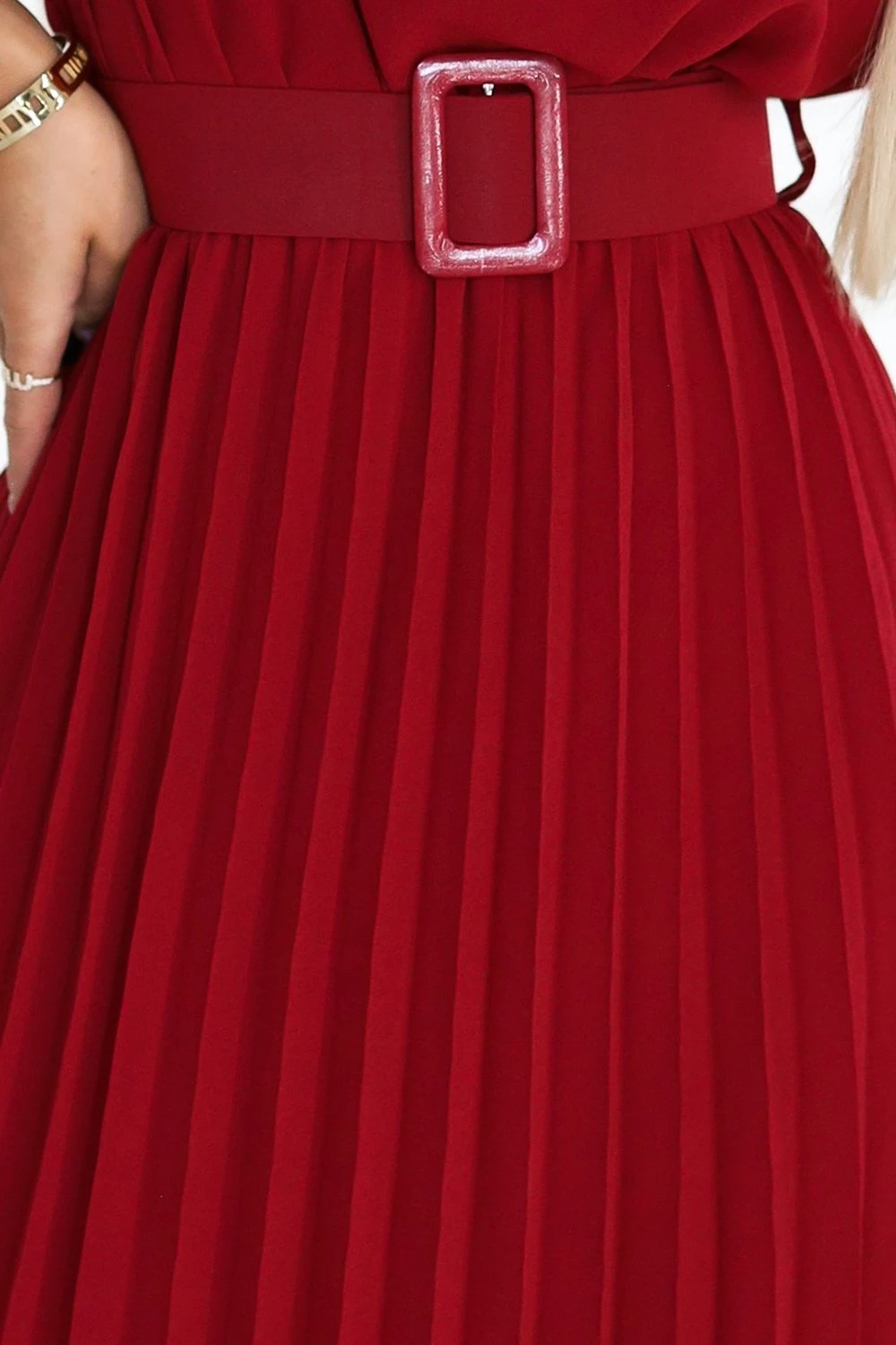 Modelka prezentuje elegancką czerwoną sukienkę plisowaną, która idealnie podkreśla sylwetkę. Sukienka plisowana w kolorze czerwonym jest uzupełniona stylowym paskiem z ozdobną klamrą, dodającym całości szyku.