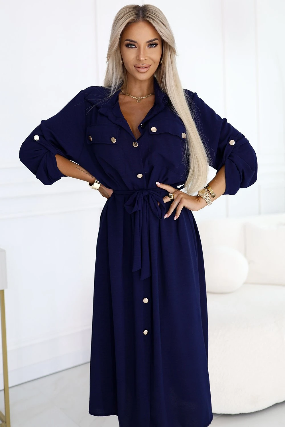 Kobieta na zdjęciu prezentuje elegancką sukienkę koszulową w głębokim granatowym kolorze, która idealnie podkreśla jej sylwetkę. Sukienka koszulowa z ozdobnymi guzikami i wiązaniem w talii dodaje stylu i klasy każdej stylizacji.