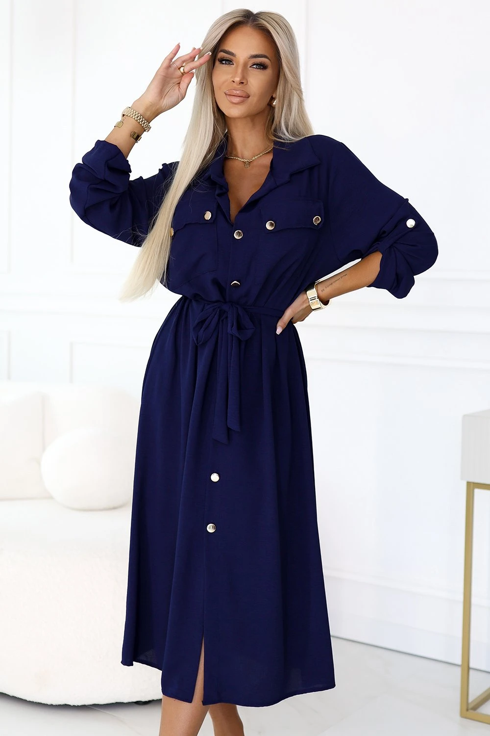 Modelka prezentuje elegancką sukienkę koszulową, która wyróżnia się głębokim granatowym kolorem i ozdobnymi guzikami. Sukienka koszulowa z wiązaniem w talii podkreśla sylwetkę, dodając stylizacji szyku i klasy.