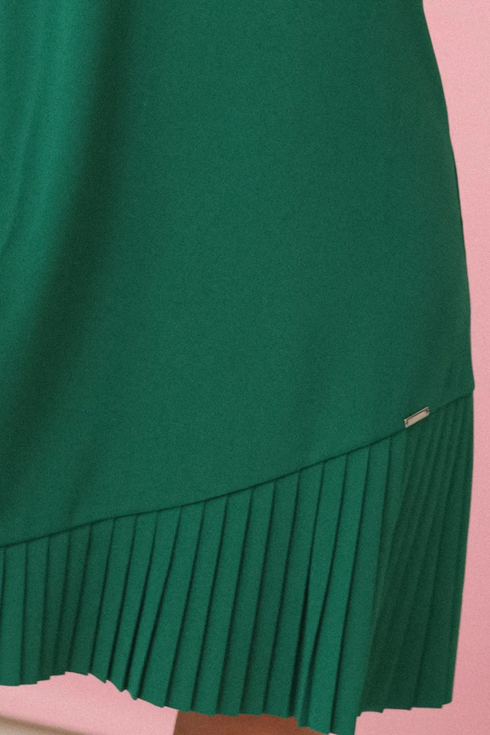Modelka prezentuje elegancką zieloną sukienkę z plisowanym dołem, która dodaje stylizacji lekkości i dynamiki. Ta zielona sukienka jest idealnym wyborem na każdą okazję, łącząc klasykę z nowoczesnym akcentem.