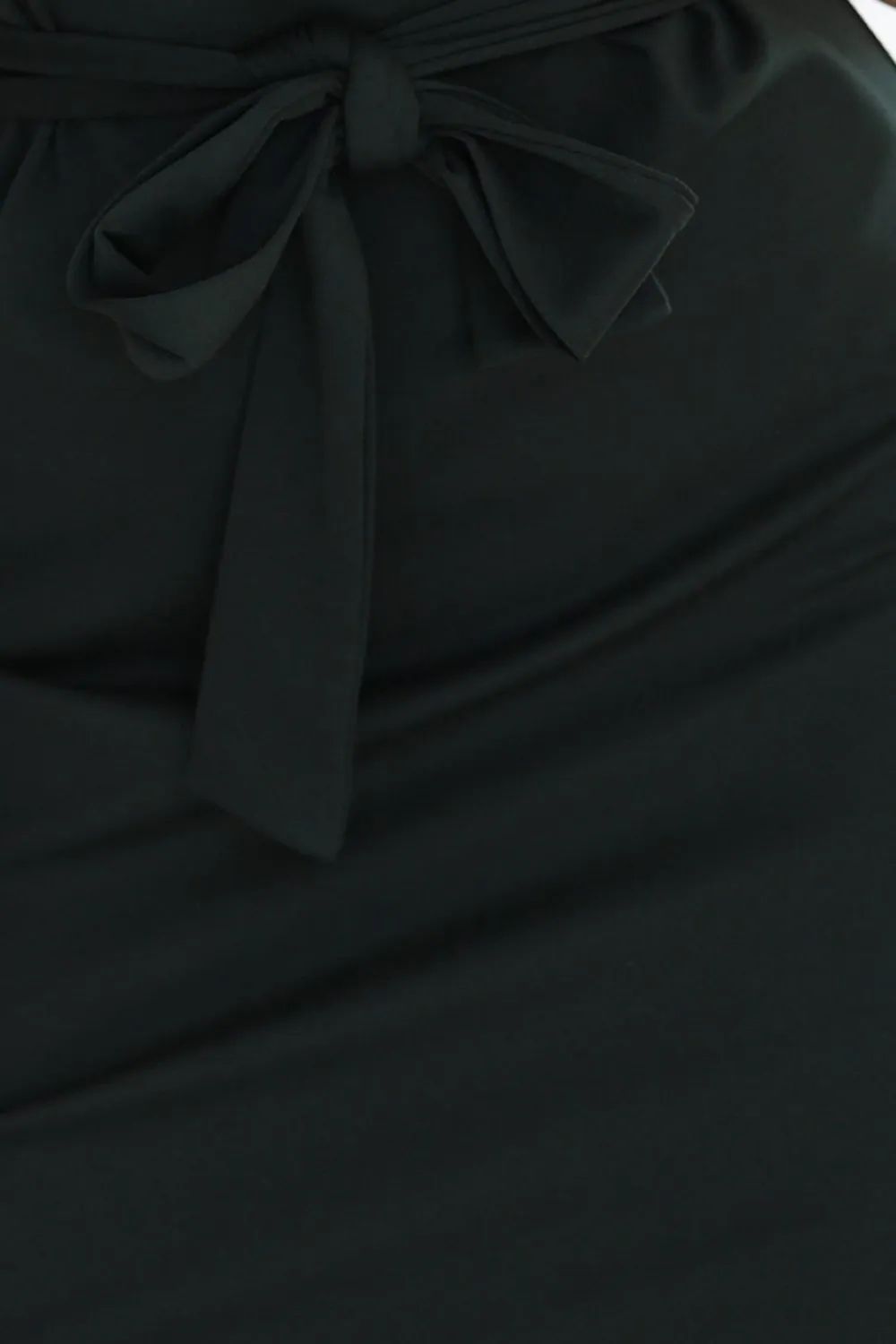 Modelka prezentuje elegancką czarną sukienkę z ozdobnym wiązaniem w talii, która podkreśla sylwetkę. Ta sukienka z kokardą dodaje stylizacji subtelnej elegancji i klasy.