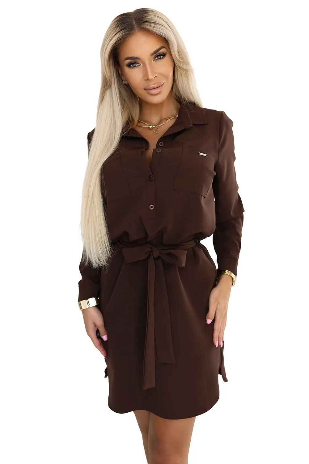 Kobieta na zdjęciu prezentuje elegancką sukienkę koszulową w głębokim odcieniu brązu, która idealnie podkreśla sylwetkę dzięki wiązaniu w talii. Sukienka koszulowa z długim rękawem i klasycznym kołnierzykiem to doskonały wybór na każdą okazję, łącząc styl z wygodą.