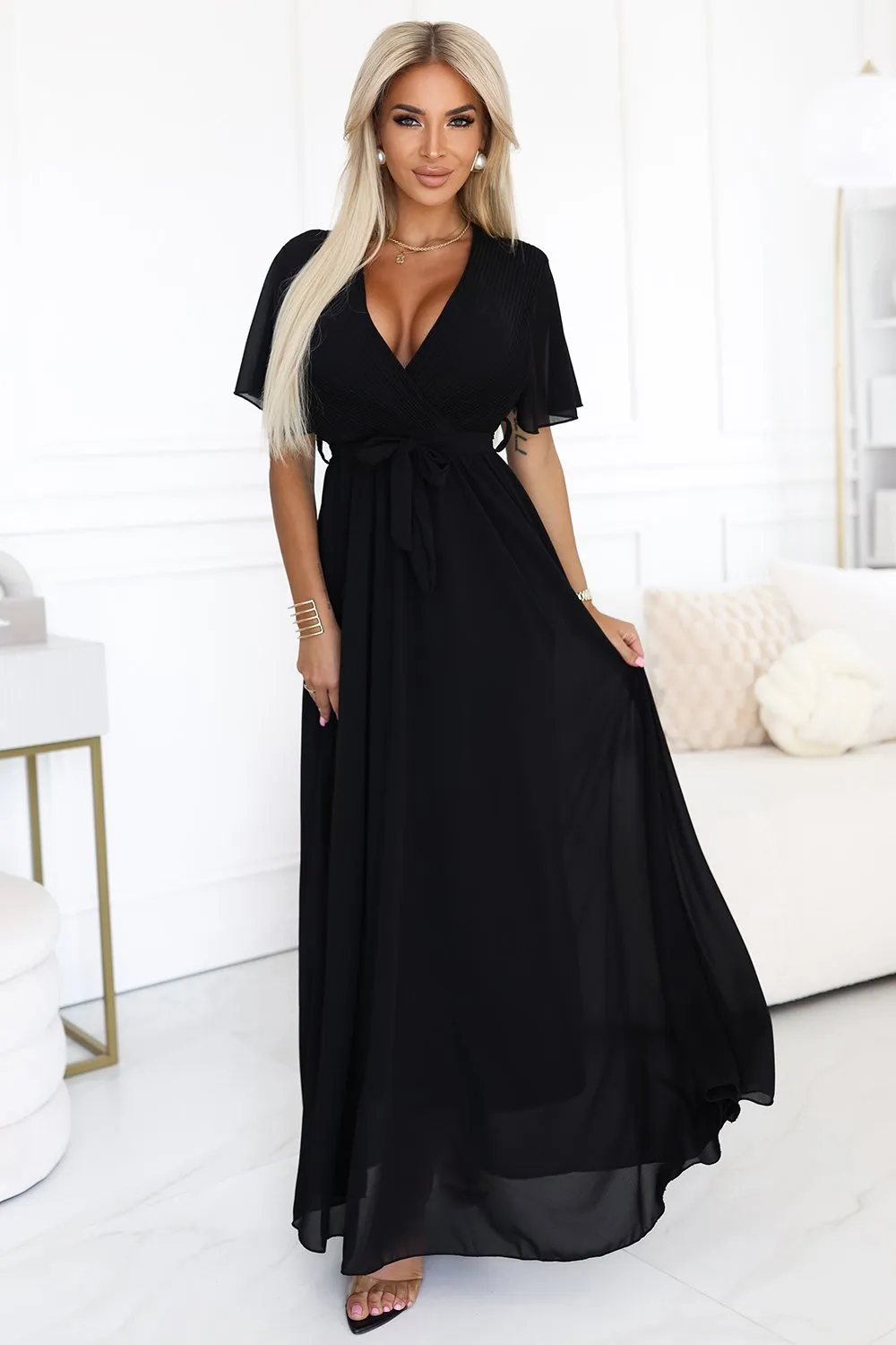 Kobieta na zdjęciu prezentuje elegancką sukienkę maxi w kolorze czarnym, która podkreśla jej sylwetkę i dodaje szyku. Sukienka maxi z wiązaniem w talii idealnie nadaje się na wieczorne wyjścia, łącząc klasykę z nowoczesnym stylem.