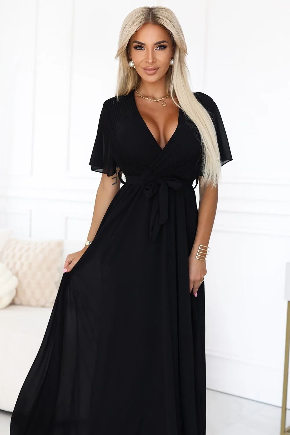 Kobieta na zdjęciu prezentuje elegancką sukienkę maxi, która podkreśla jej sylwetkę i dodaje klasy każdej stylizacji. Ta czarna sukienka maxi z wiązaniem w talii to idealny wybór na wieczorne wyjścia, łącząc w sobie szyk i wygodę.