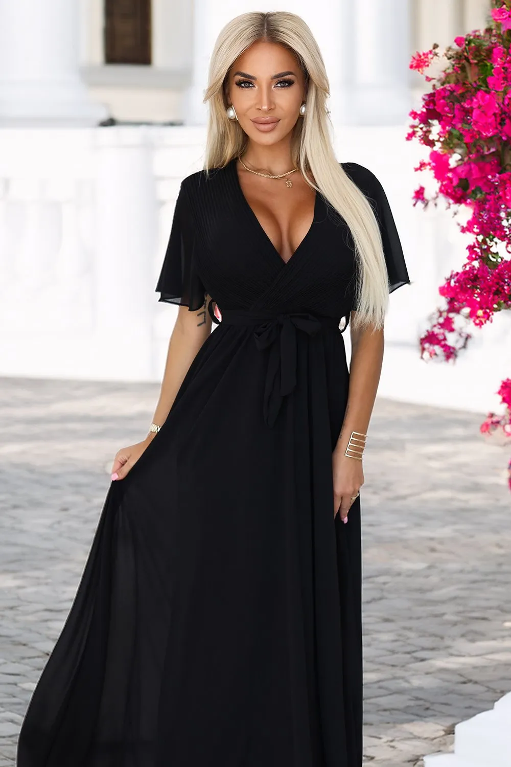 Modelka prezentuje elegancką czarną sukienkę maxi, która podkreśla sylwetkę i dodaje szyku. Sukienka maxi z głębokim dekoltem i delikatnym paskiem w talii to idealny wybór na wieczorne wyjścia.