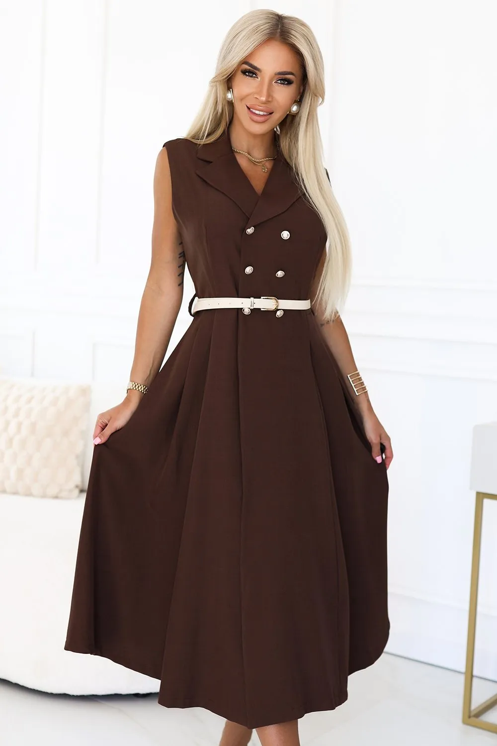 Kobieta na zdjęciu prezentuje elegancką sukienkę bez rękawów w kolorze czekoladowym, która podkreśla jej sylwetkę. Sukienka jest ozdobiona kontrastowym paskiem i stylowymi guzikami, co dodaje jej klasycznego uroku.