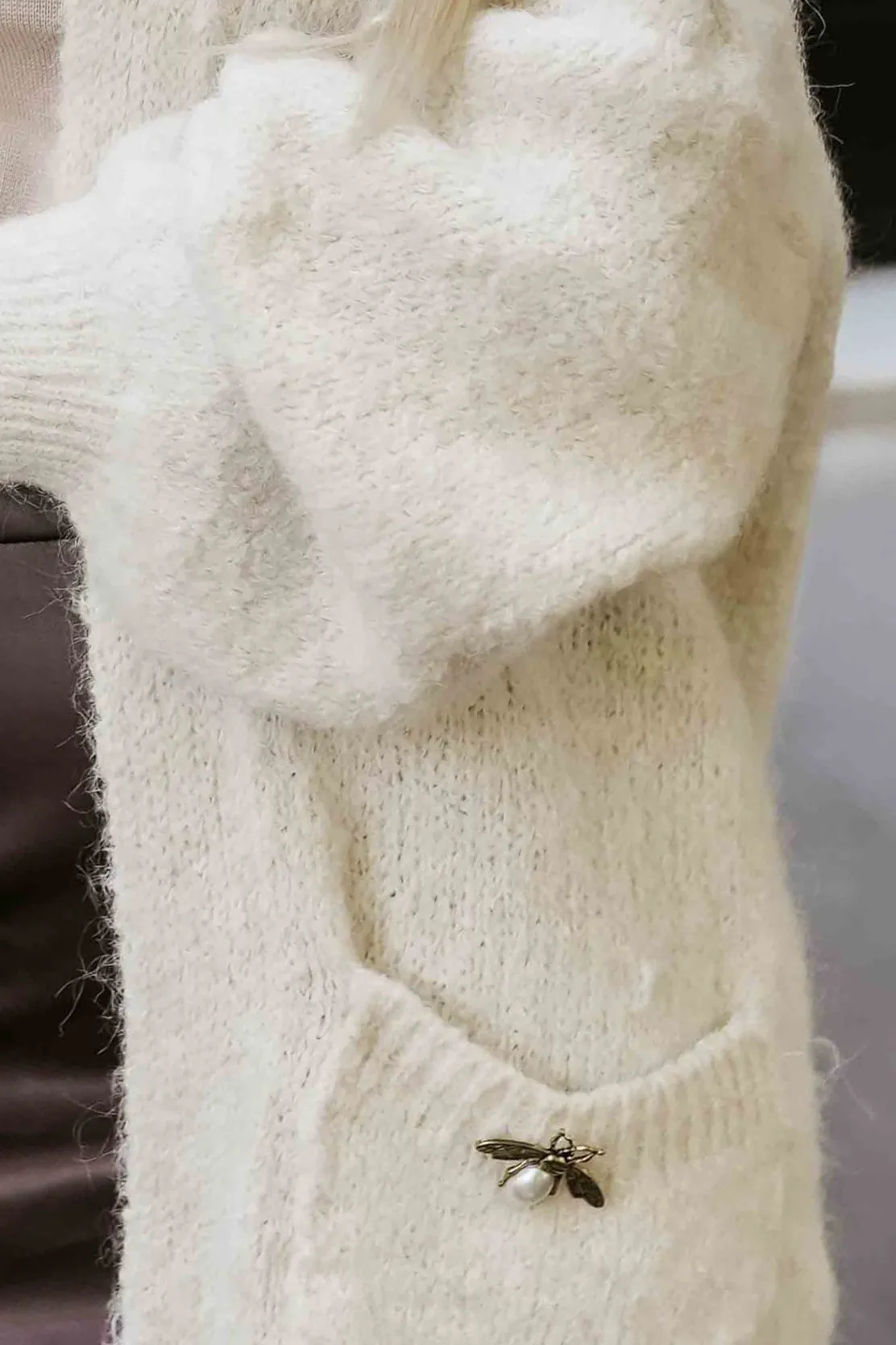 Modelka prezentuje elegancki sweter z delikatnej dzianiny, który wyróżnia się subtelną broszką w kształcie pszczoły. Sweter z dzianiny dodaje stylizacji klasy i wyjątkowego charakteru, idealnie wpisując się w trendy sezonu.