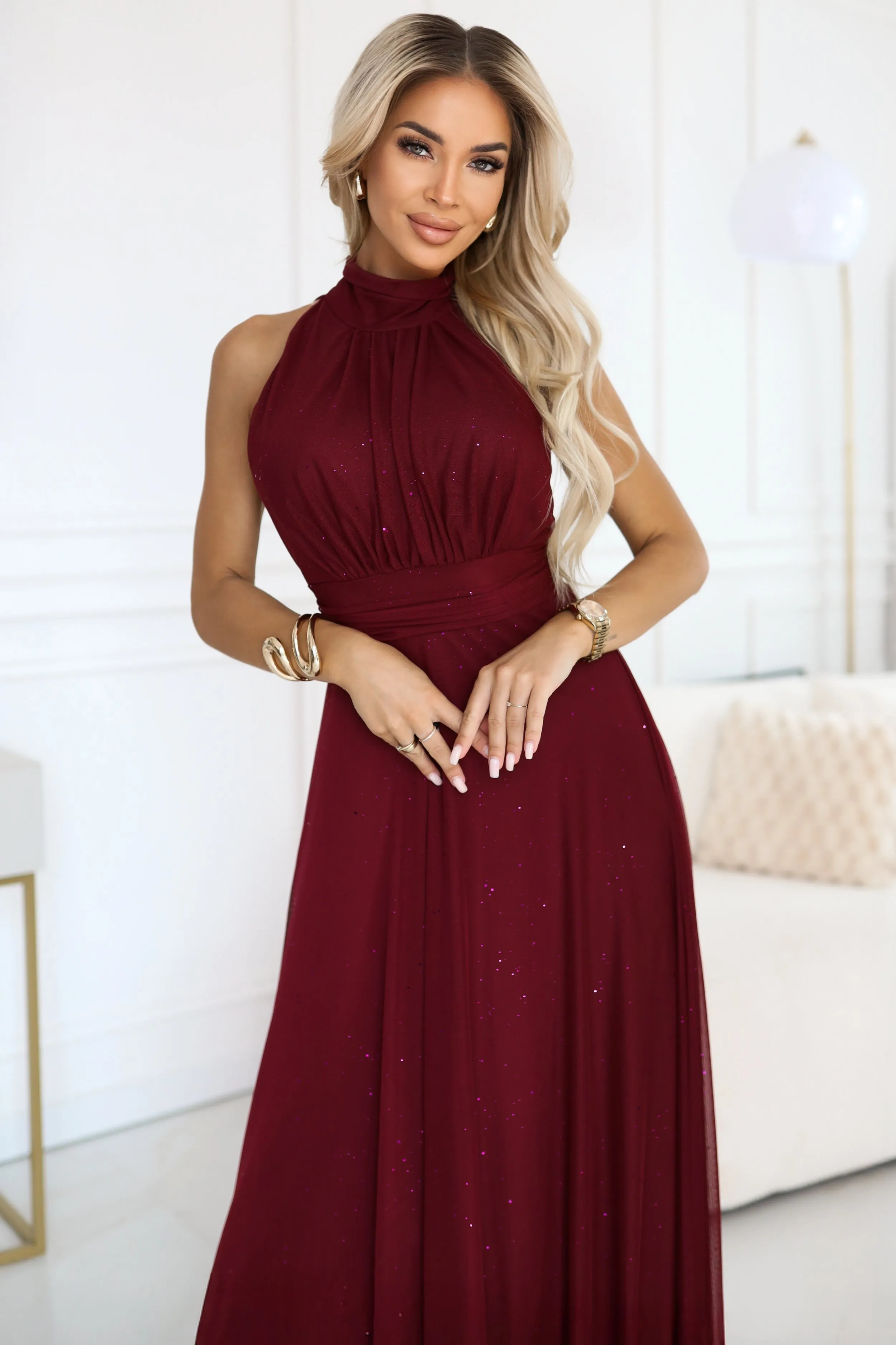 Modelka prezentuje elegancką sukienkę wieczorową w odcieniu głębokiego bordo, którą cechuje subtelny połysk. Sukienka wieczorowa świetnie podkreśla sylwetkę dzięki swojej dopasowanej górze i zwiewnemu dołowi.